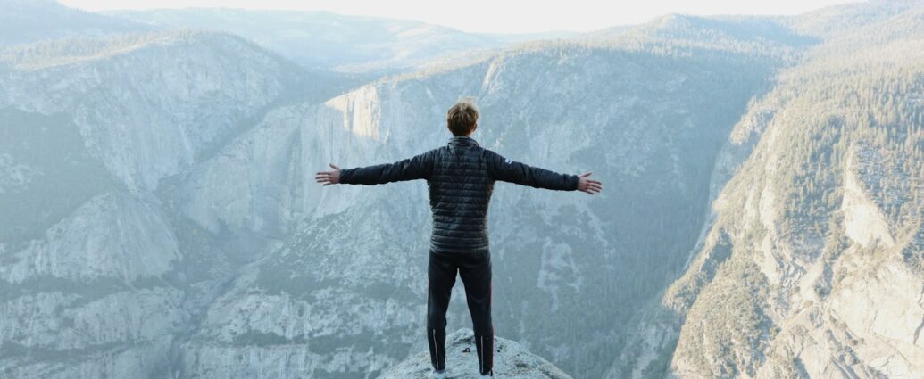 man opening his arms wide open on snow covered cliff with view of mountains during daytime