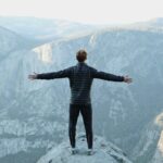 man opening his arms wide open on snow covered cliff with view of mountains during daytime